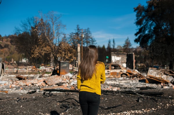 A woman in yellow sweater stands in front of her burned down home