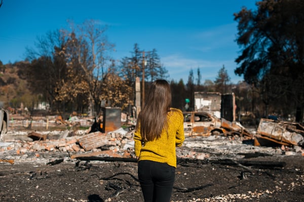 A woman in yellow sweater stands in front of her burned down home